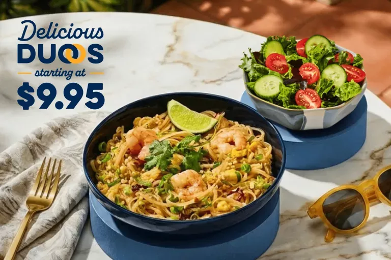 Delicious Duos starting at $9.95. Pad Thai with Shrimp and Side Garden Salad on a marble patio table with a fork, napkin and sunglasses