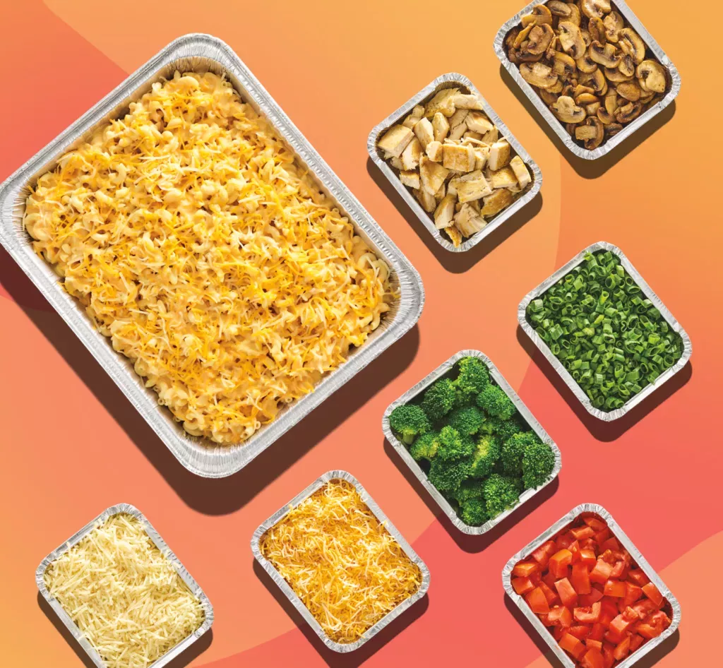 Tray of macaroni and cheese with various toppings that include but are not limited to grilled chicken, mushrooms, green onions, broccoli and tomatoes.
