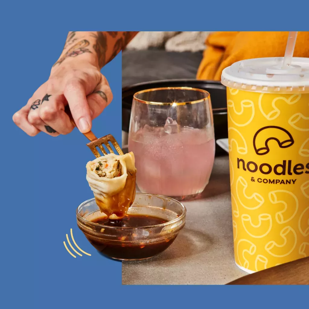 Hand dipping a potsticker on a fork into a bowl of potsticker sauce, next to a pink drink in a clear glass and a yellow fountain drink cup.