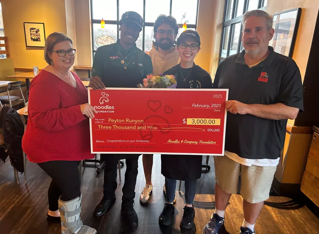 Noodles and Company 2025 Scholarship Award Winner holding a large check surrounded by friends, family and fellow employees.