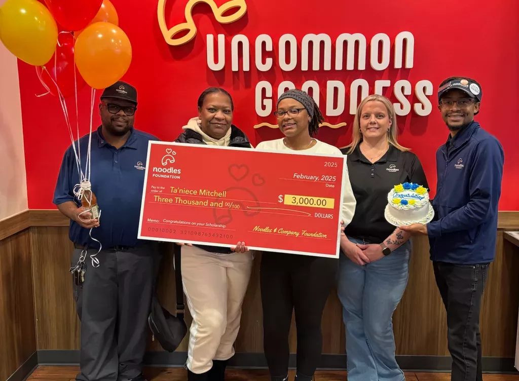 Noodles and Company 2025 Scholarship Award Winner holding a large check surrounded by friends, family and fellow employees.