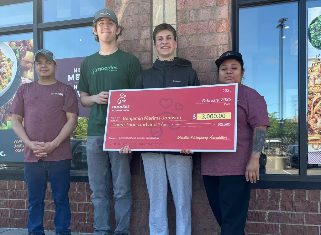 Noodles and Company 2025 Scholarship Award Winner holding a large check surrounded by friends, family and fellow employees.