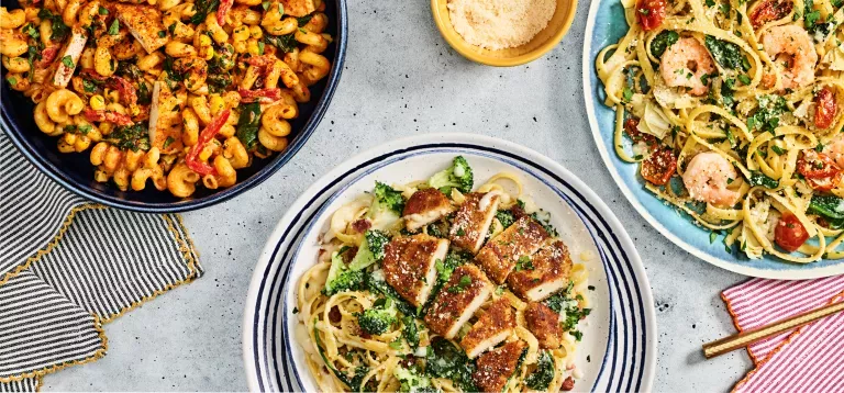 Noodles &amp; Company's Chipotle Chicken Cavatappi, Crispy Chicken Bacon Alfredo, and Lemon Garlic Shrimp Scampi on decorative plates with a side of grate parmesan and utensils.