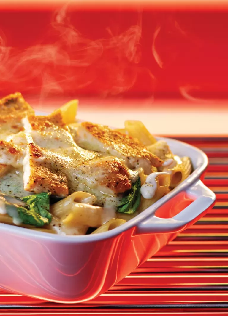 Baked Alfredo with grilled chicken, penne noodles, cheese, and spinach in a white dish with steam rising from the food.