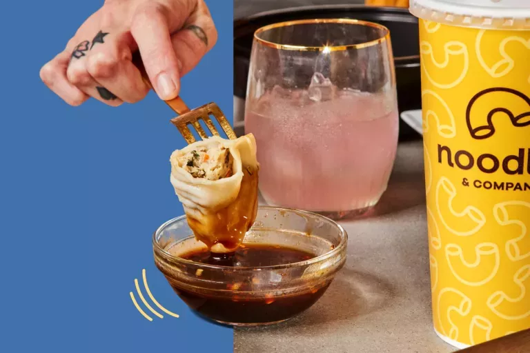 Hand dipping a potsticker on a fork into a bowl of potsticker sauce, next to a pink drink in a clear glass and a yellow fountain drink cup.