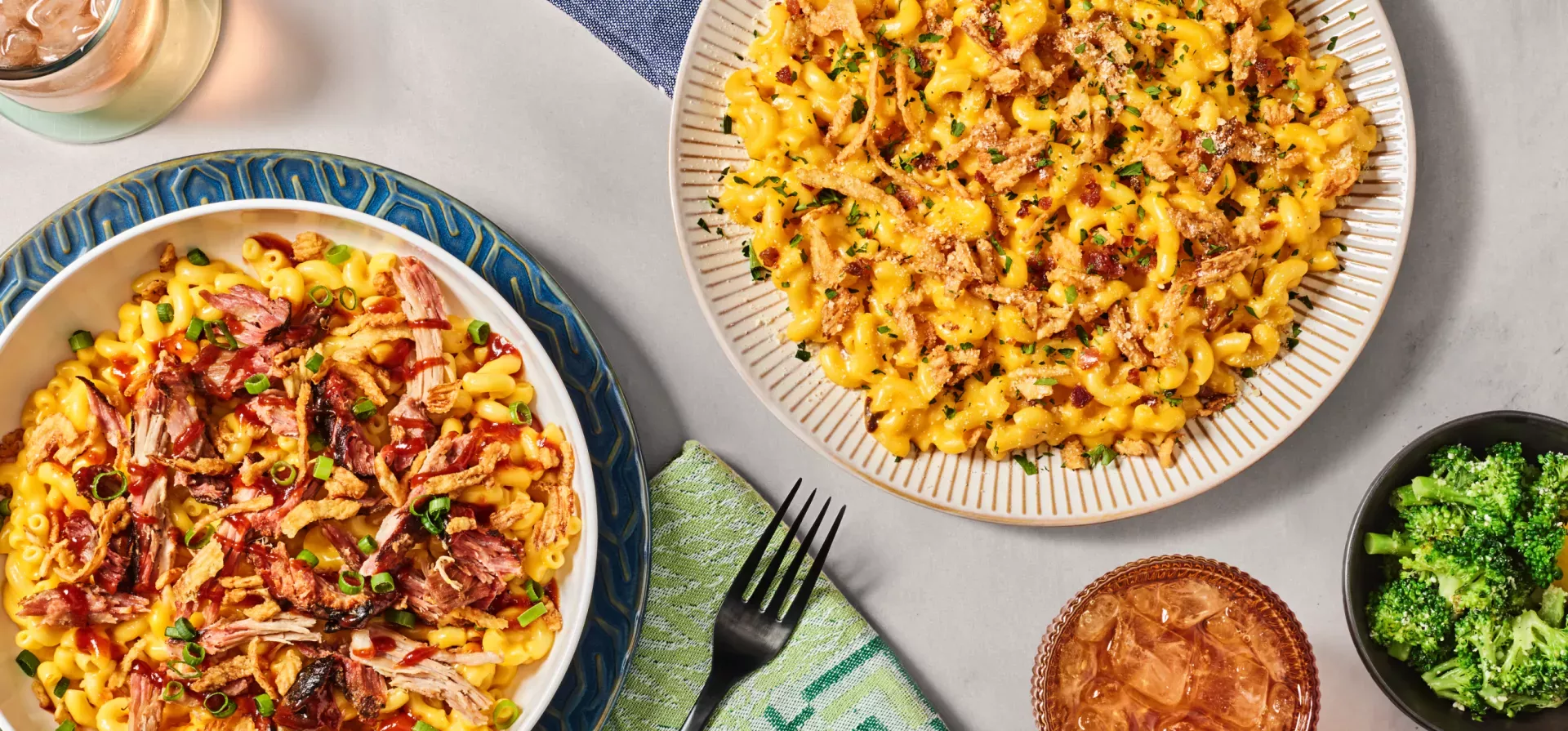 Tabletop image of pulled pork bbq mac, garlic bacon crunch mac an iced drink and a side of broccoli